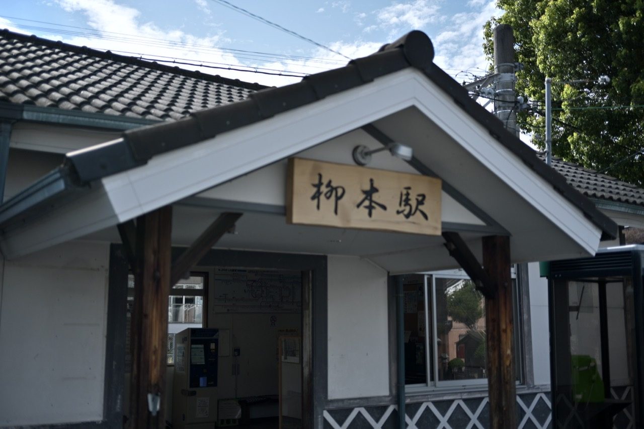 黒い瓦屋根と白い壁の柳本駅舎に、柳本駅の木製看板が掲げられている