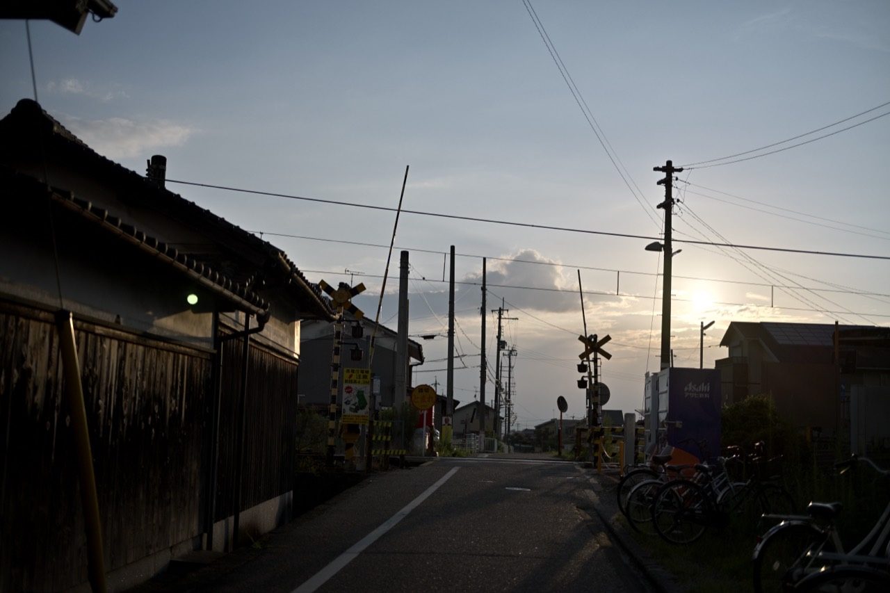 傾いた夕日を背に、踏切と自転車、木造の家並みが影になる